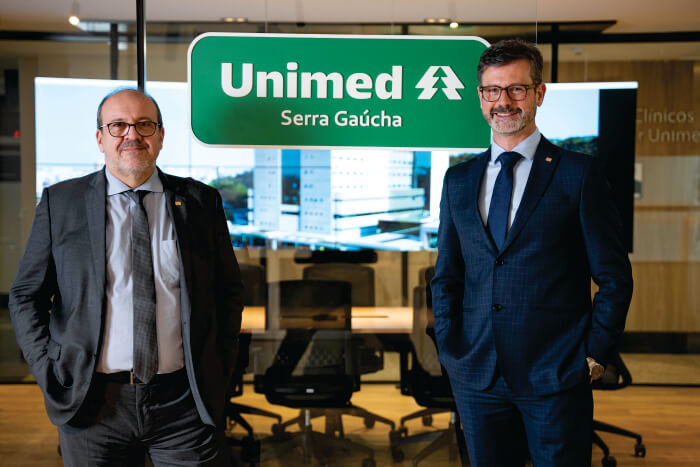 Dois homens sorrindo em frente à logo da Unimed, verde com fonte branca